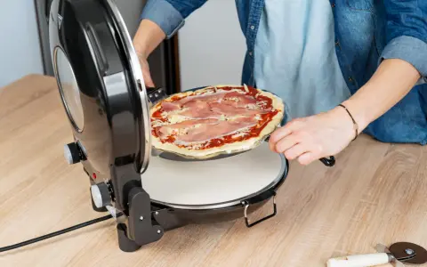 Osoba wkłada surowe ciasto na pizzę z szynką do okrągłego piekarnika.