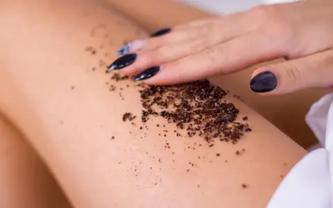 Dłoń z czarnym lakierem rozprowadza na skórze odżywczy peeling z kawy, przygotowując ją na letnie słońce.