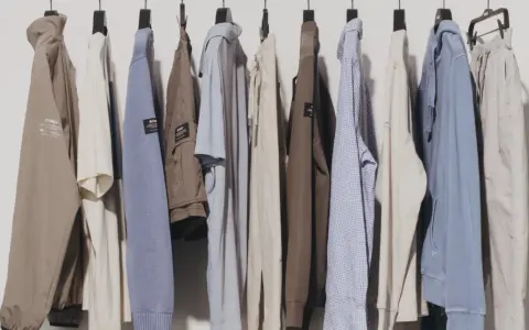 Eine Reihe von Kleidungsstücken, darunter Jacken, Pullover und Hosen, hängen an Kleiderbügeln. Diese zeitlose Kleidung in gedeckten Farben strahlt Komfort und Stil aus.