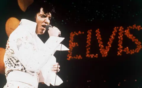 Elvis Presley singt in seinem berühmten weißen Anzug in Las Vegas, während sein Name in roten Lichtern erstrahlt.