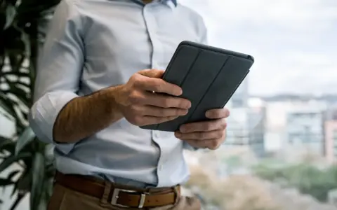 Mężczyzna w jasnej koszuli i brązowych spodniach trzyma tablet w szarej okładce. e cmr.