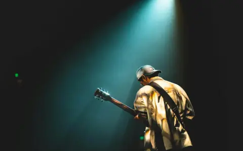Mężczyzna w czapce z daszkiem gra na gitarze pod światłem reflektora, tworząc atmosferę jak z najsłynniejszych utworów rockowych.