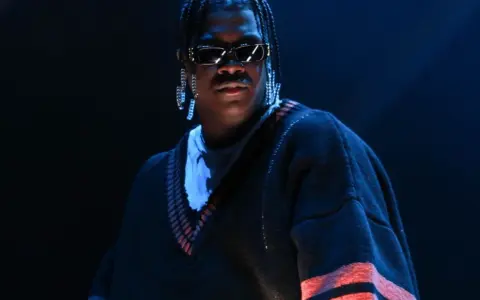 Lil Yachty w okularach i długich kolczykach, w swetrze z pomarańczowymi pasami. Let's start here.