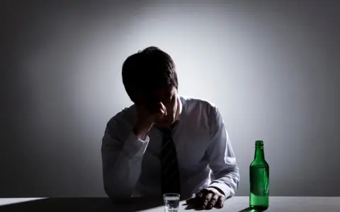 Mężczyzna w białej koszuli i krawacie siedzi przy stole, trzymając się za głowę. Obok stoi szklanka i zielona butelka. To obraz przedawkowania alkoholu.