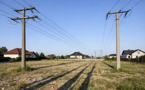 Linia wysokiego napięcia nad działką z rozpoczętą budową domu. Słupy energetyczne i przewody przecinają krajobraz z kilkoma domami w tle.