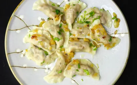Pyszne pierogi z kaszą gryczaną, okraszone skwarkami i posypane natką pietruszki, na talerzu z delikatnym wzorem kwiatowym.