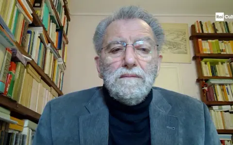 Uomo anziano con barba grigia, occhiali e giacca, circondato da scaffali pieni di libri. Il suo sguardo intenso suggerisce che ei fu significato.