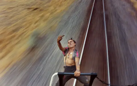 A defiant young man on a train, captured by mike brodie photography, flips the bird against a blur of motion.
