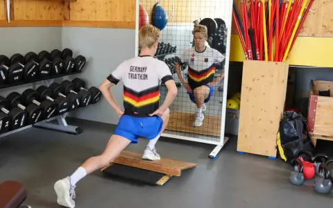 Ein Athlet im "Germany Triathlon"-Shirt macht Ausfallschritte im Athletik Training. Hanteln und Trainingsgeräte im Hintergrund.