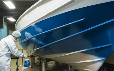 Un peintre en combinaison blanche applique une peinture bleue brillante sur la coque d'un bateau dans un atelier.