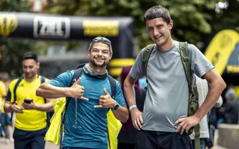 Zwei Männer mit Rucksäcken, einer mit Daumen hoch, der andere mit Handzeichen. Sie sind bereit für ihr Abenteuer, bei dem die richtige **Merinowolle Pflege** wichtig ist.