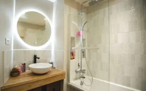 Une **salle de bain blanche** moderne avec un lavabo rond sur un meuble en bois, un miroir lumineux et une douche avec carrelage clair.