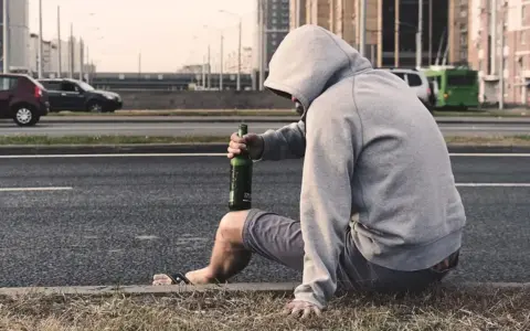Mężczyzna w kapturze siedzi przy drodze z butelką piwa. Jego postawa sugeruje walkę z nałogiem, być może potrzebuje pomocy terapeuty od uzależnień.