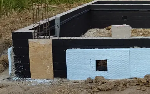 Fundament z czarną izolacją przeciwwilgociową i płytami XPS. Widać zbrojenie i fragmenty szalunku. Dobra jaka izolacja na fundament.