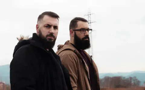 Dos hombres con barba posan con seriedad. Uno viste chaqueta negra, el otro marrón. Parecen listos para un rap de "falsa alarma".