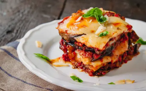 Una fetta di parmigiana di melanzane al forno, stratificata con melanzane, sugo e formaggio filante, guarnita con basilico fresco.