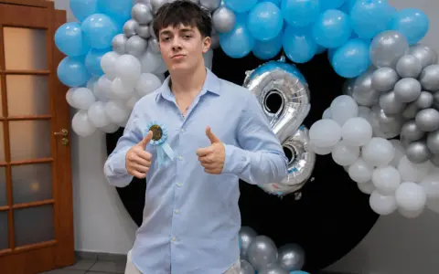 Chłopak w błękitnej koszuli z rozetką "18th Birthday" i balonami.