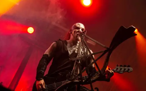 Un musicien au visage peint, jouant de la basse sur scène, baigné de lumières rouges. Sa posture exprime une vie d'agonie.