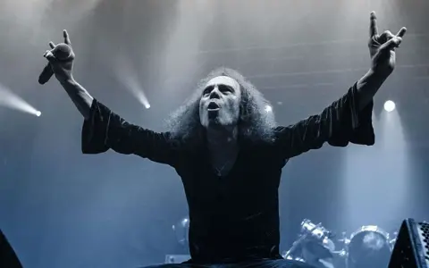 Ronnie Dio mit erhobenen Armen und Mikrofon, die Hand zum "Rock on"-Gruß geformt.