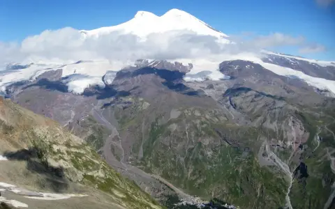 Majestatyczny Elbrus, najwyższy szczyt Europy, góruje nad skalistymi zboczami i lodowcami. W dole widać niewielką osadę, która pokazuje, gdzie leży Elbrus.