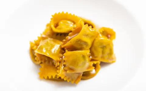 Un mucchietto di ravioli del plin, dorati e conditi con un sugo cremoso, su un piatto bianco.