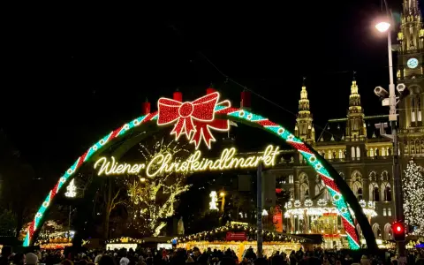 Wiedeński jarmark świąteczny w blasku świateł. Tłumy ludzi spacerują pod łukiem z napisem "Wiener Christkindlmarkt", w tle widać zabytkowe budynki i choinkę.