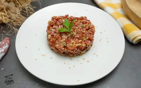 Tartar de fuet picado finamente, con semillas de sésamo y perejil, servido en plato blanco.