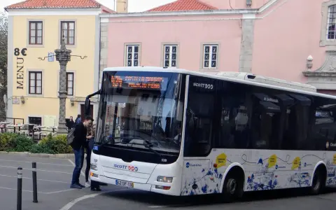 Ein Scott-Bus in Portugal hält an einer Haltestelle. Touristen steigen ein, während im Hintergrund bunte Gebäude und ein historischer Brunnen zu sehen sind.