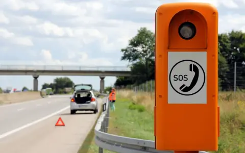 Ihr Fahrzeug ist auf der Autobahn liegen geblieben. Eine Frau in Warnweste steht neben dem offenen Kofferraum, ein Warndreieck liegt auf der Fahrbahn. Eine Notrufsäule steht im Vordergrund.