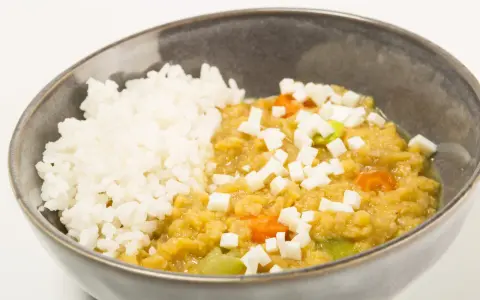 Un tazón de arroz blanco junto a un guiso cremoso de lentejas rojas con trozos de queso y verduras.