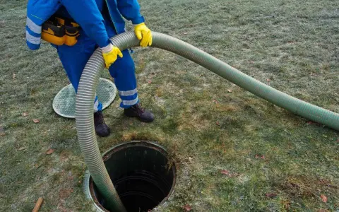 Pracownik w niebieskim kombinezonie i żółtych rękawicach wykonuje czyszczenie szamba, wkładając gruby wąż do otwartego włazu.