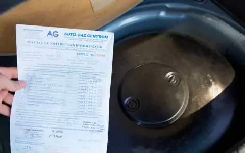Fragment dokumentu z danymi homologacji butli LPG obok okrągłej pokrywy w bagażniku samochodu.