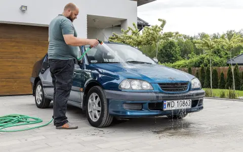 Mężczyzna myje niebieskie auto pod domem w upalny dzień. Wąż ogrodowy leży na podjeździe.