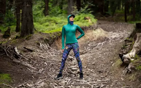 Kobieta w lesie, w kolorowych legginsach i zielonej bluzie, pokazuje, co daje stretching: elastyczność i siłę do pokonywania górskich ścieżek.