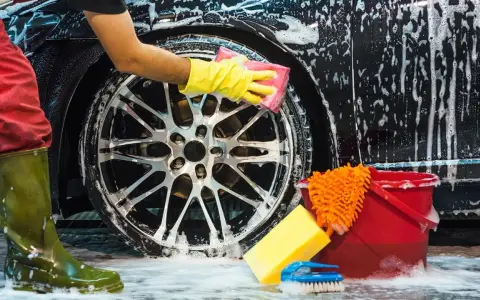 Persona con guantes amarillos detallando la llanta de un coche negro con espuma, mostrando cómo lavar el coche a mano. Cubo rojo y esponjas cerca.