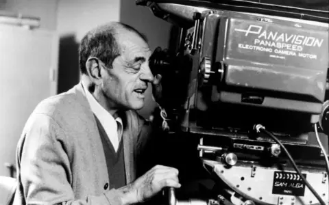 Un regista spagnolo, con un maglione grigio, guarda attraverso l'obiettivo di una cinepresa Panavision.