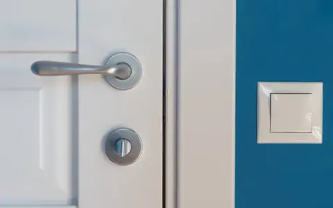 Eleganckie, metalowe klamki do białych drzwi idealnie komponują się z niebieską ścianą i białym włącznikiem.