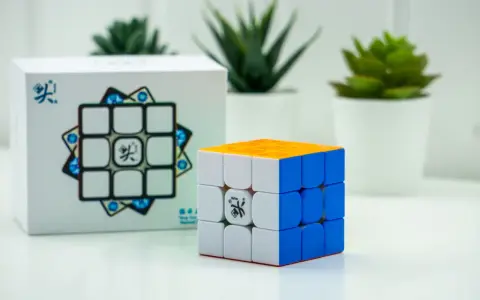 En speed cube 3x3 med orange, vit och blå sida står bredvid sin förpackning. Tre gröna växter i vita krukor syns i bakgrunden.