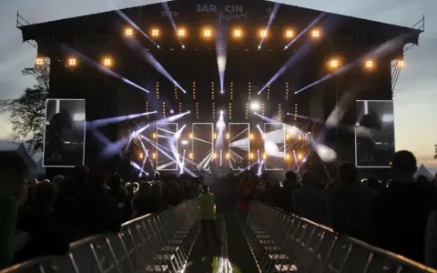 Jarocin Festiwal rozświetlony tysiącami świateł. Tłum czeka na koncert, a na scenie widać artystów.