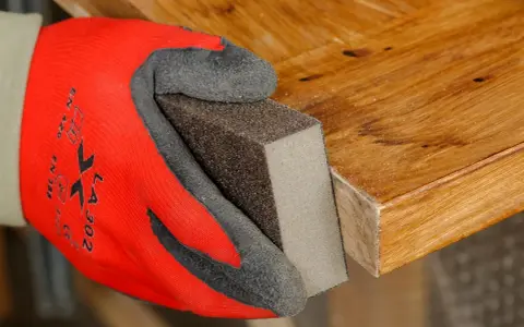 Mano con guante rojo lijando borde de puerta de madera para rebajarla.