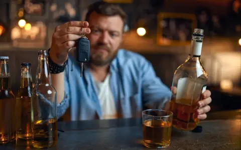 Mężczyzna trzyma kluczyki do samochodu, zastanawiając się, czy jestem pijany. Obok butelki i szklanka alkoholu.