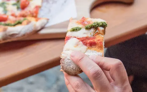 Eine Hand hält ein Stück Pizza mit Tomaten, Mozzarella und Pesto. So sieht eine köstliche Pizza aus!