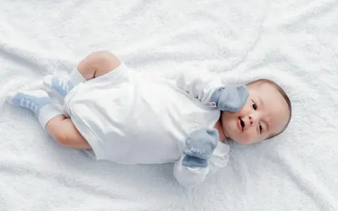 Urocze niemowlę w białym body i niebieskich skarpetkach leży na białym kocyku. Wygląda na zadowolone, choć może właśnie zastanawia się, kiedy wreszcie niemowlę nie robi kupy.