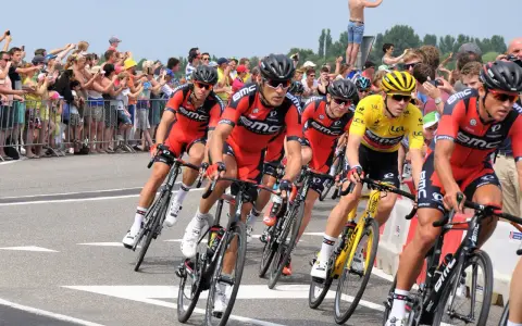 Cyclists in red BMC jerseys race, with one in yellow, as spectators cheer. A perfect scene to watch Tour de France live!