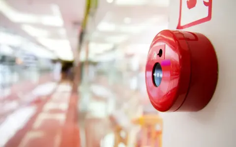 Czerwony przycisk alarmowy, kluczowy element ochrony przeciwpożarowej, zamontowany na jasnej ścianie. W tle rozmyty korytarz centrum handlowego.