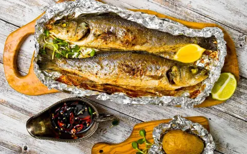 Due branzini cotti al cartoccio, dorati e succulenti, serviti con spicchi di limone, patata al cartoccio e salsa piccante.
