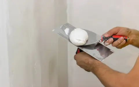 Mani che usano spatole per applicare stucco su un muro. Lo stucco serve a livellare e riparare le imperfezioni.