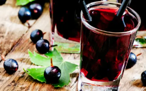 Gläser mit dunklem Johannisbeeren Schnaps, umgeben von frischen Beeren und Blättern auf Holz.