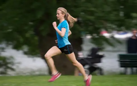 En person som springer snabbt över en grön gräsmatta, med träd och en sjö i bakgrunden.
