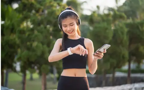 Lächelnde junge Frau mit Kopfhörern und Smartphone, die ihre Smartwatch prüft. Sie trägt atmungsaktive Kleidung für ihr Training im Freien.